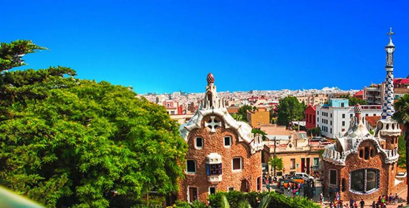 Vue générale du quartier de l'Eixample à Barcelone, avec des toits d'immeubles modernistes et la Sagrada Familia à l'horizon, sous un ciel bleu.