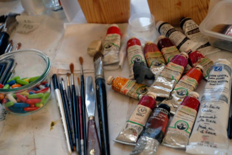 Gros plan sur un pinceau d'aquarelle trempé dans la peinture, prêt à déposer la couleur sur un papier texturé, avec des pots d'eau et des couleurs en arrière-plan.