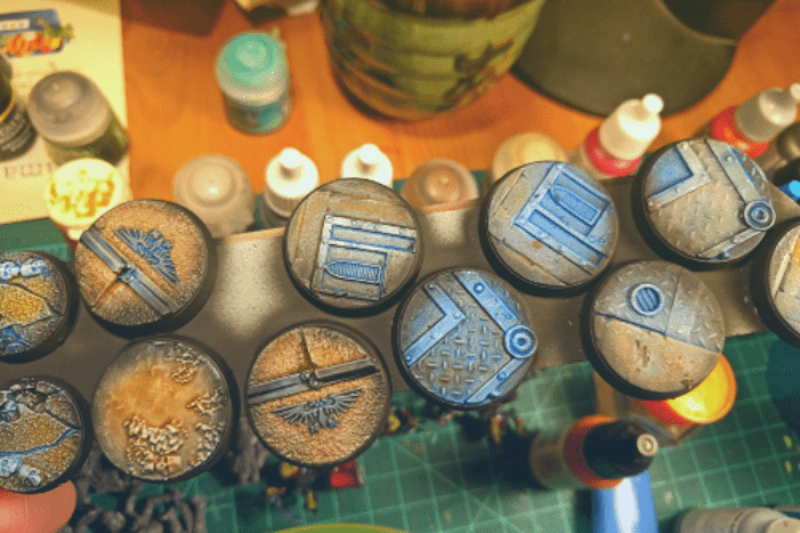 Figurine peinte posée sur un socle détaillé représentant un sol rocheux avec de la végétation statique et des graviers.